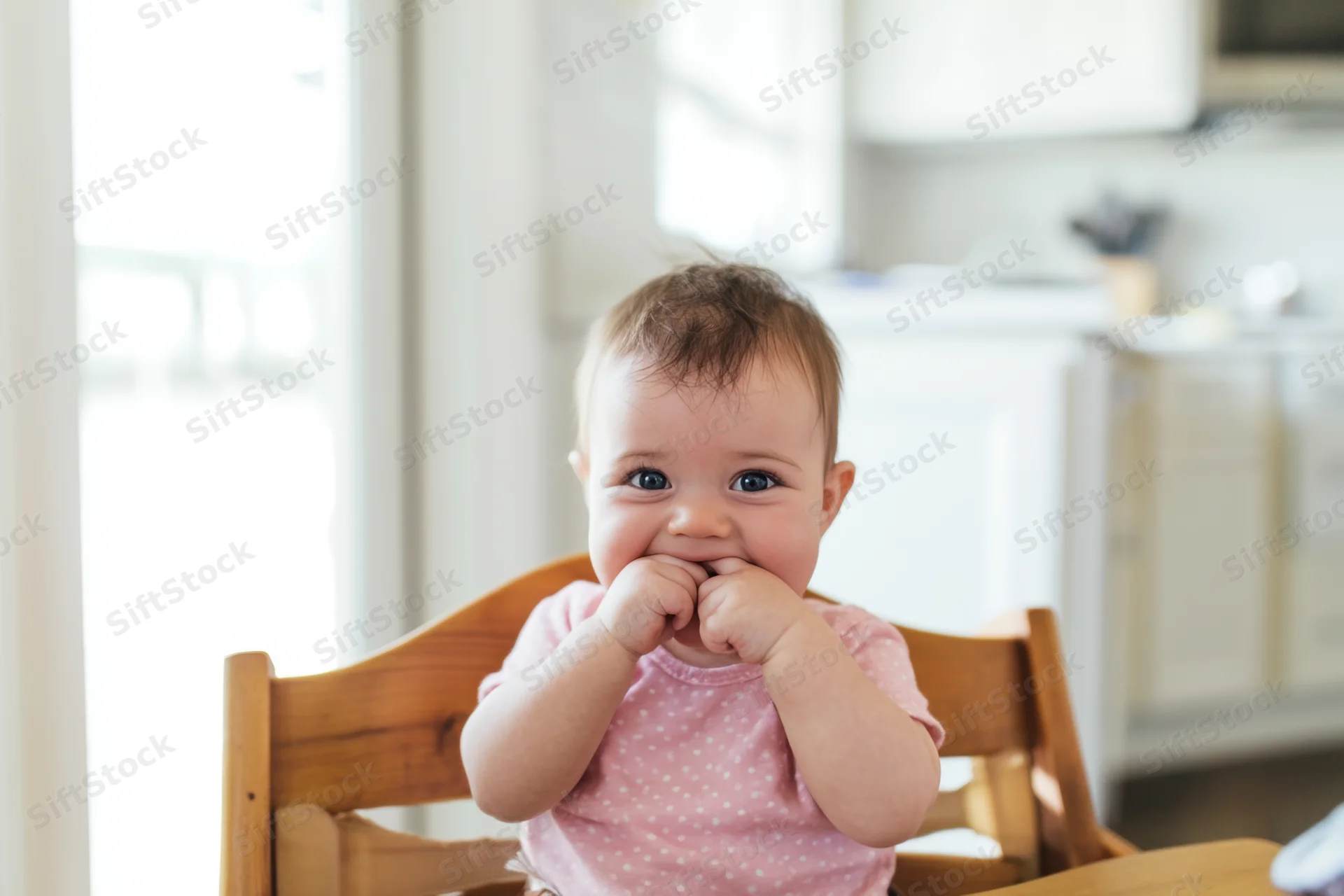 Baby sitting in wooden high chair in bright kitchen chewing fingers