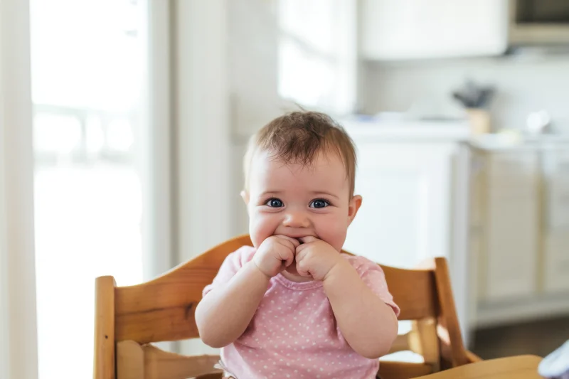 Baby sitting in wooden high chair in bright kitchen chewing fingers