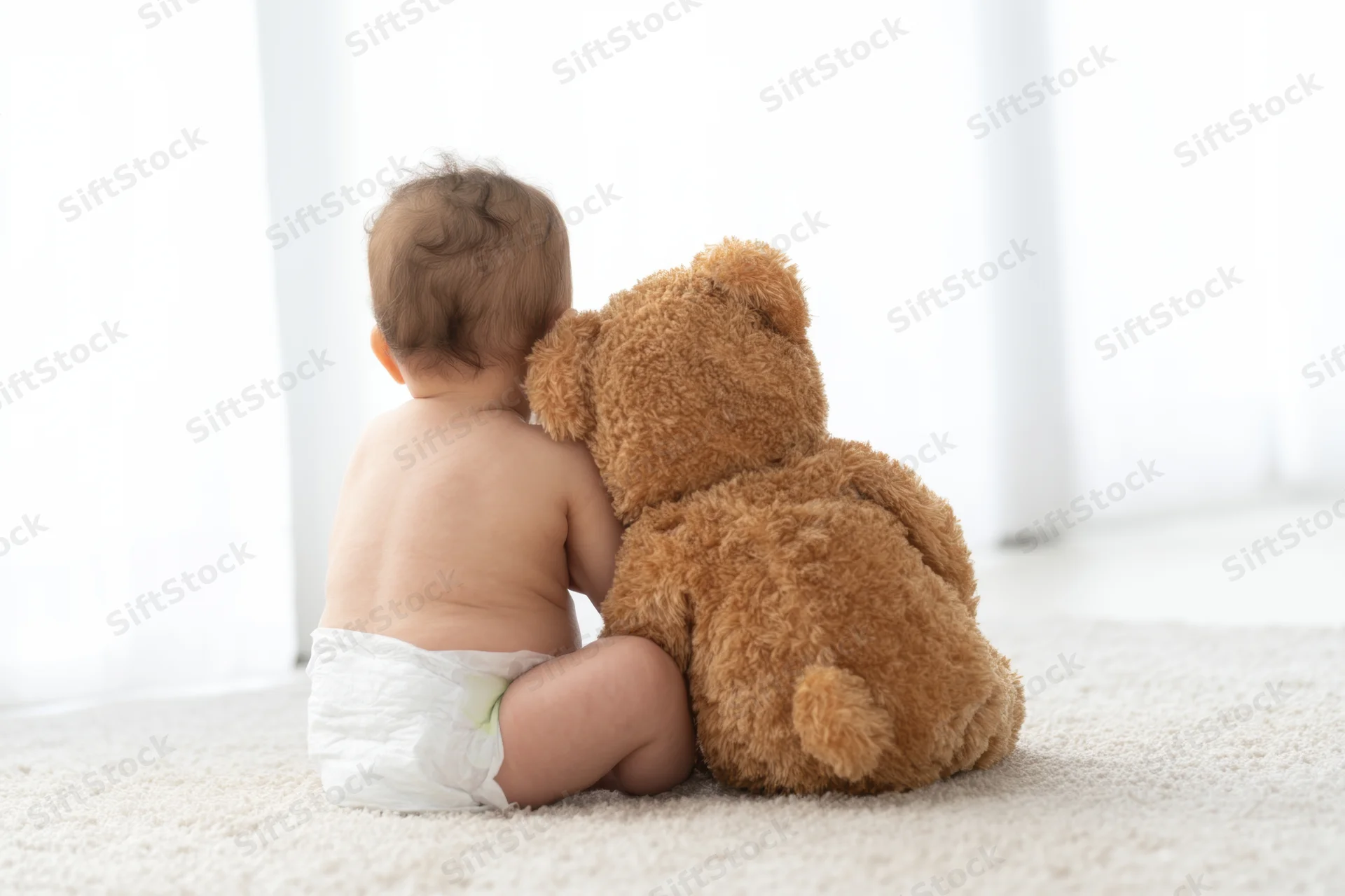 Baby sitting on light carpet seen from behind hugging a brown teddy bear