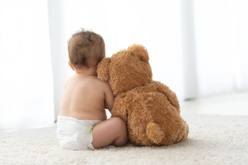 Baby sitting on light carpet seen from behind hugging a brown teddy bear
