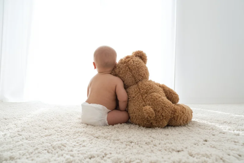 Baby sitting on light carpet seen from behind with teddy bear, wearing diaper