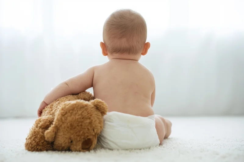 Baby sitting on light carpet seen from behind with teddy bear, wearing diaper