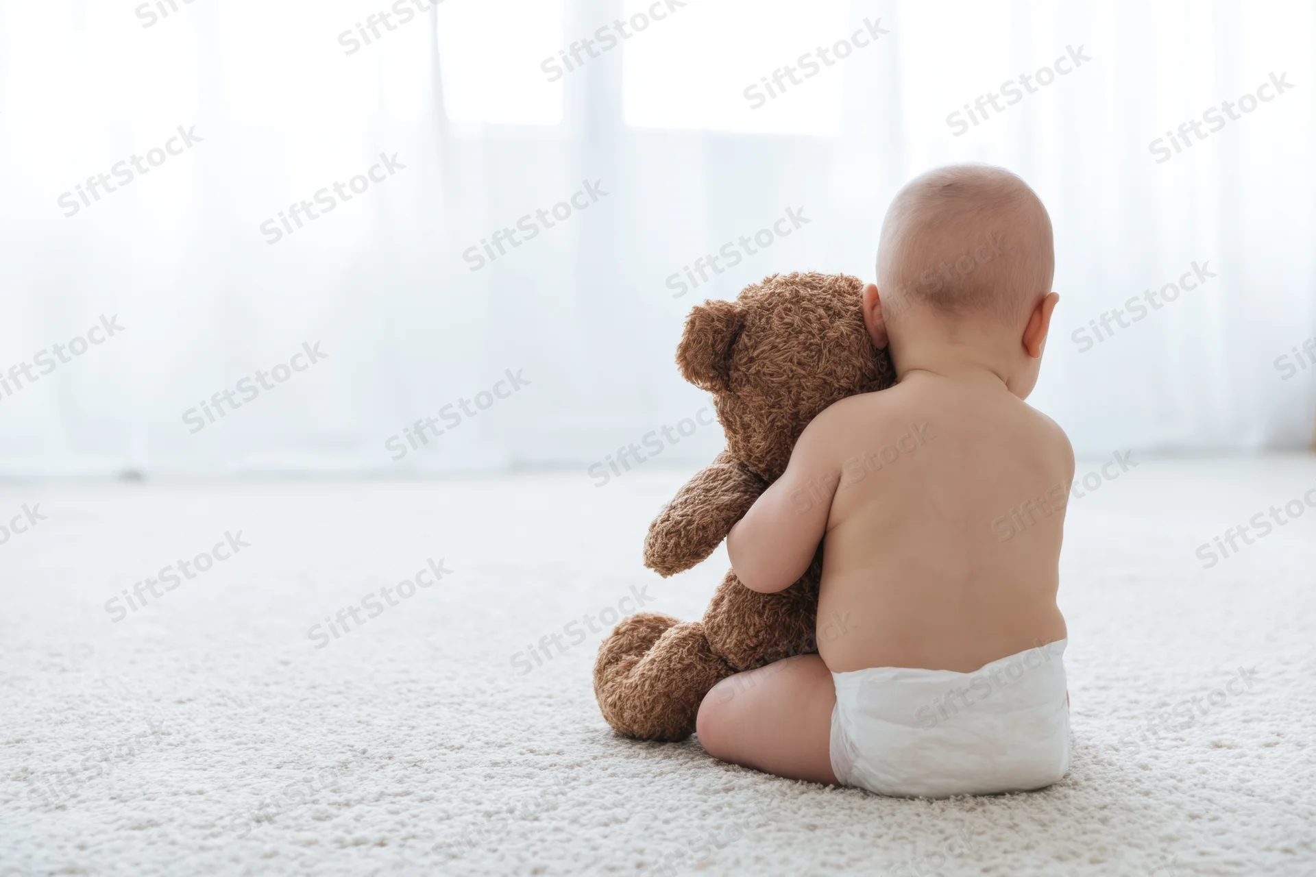 Baby sitting on light carpet seen from behind hugging a brown teddy bear wearing a diaper