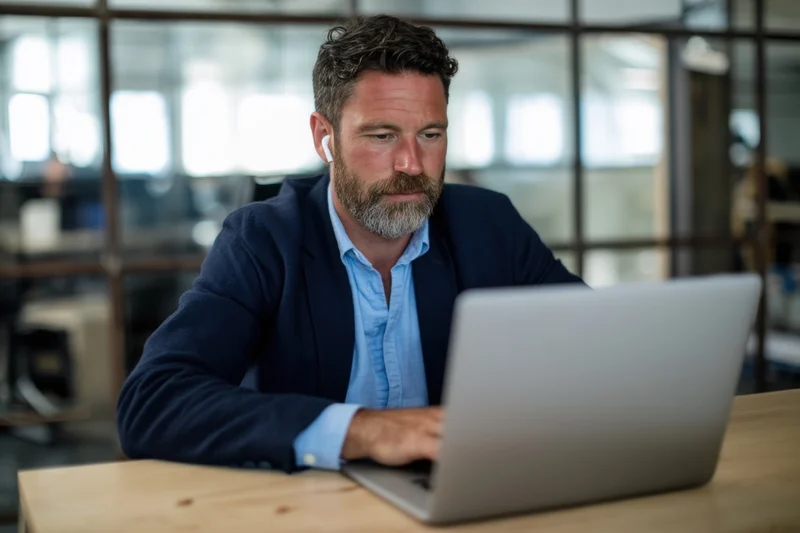Bearded man in navy blazer and light blue shirt working on laptop at wooden desk in modern office space