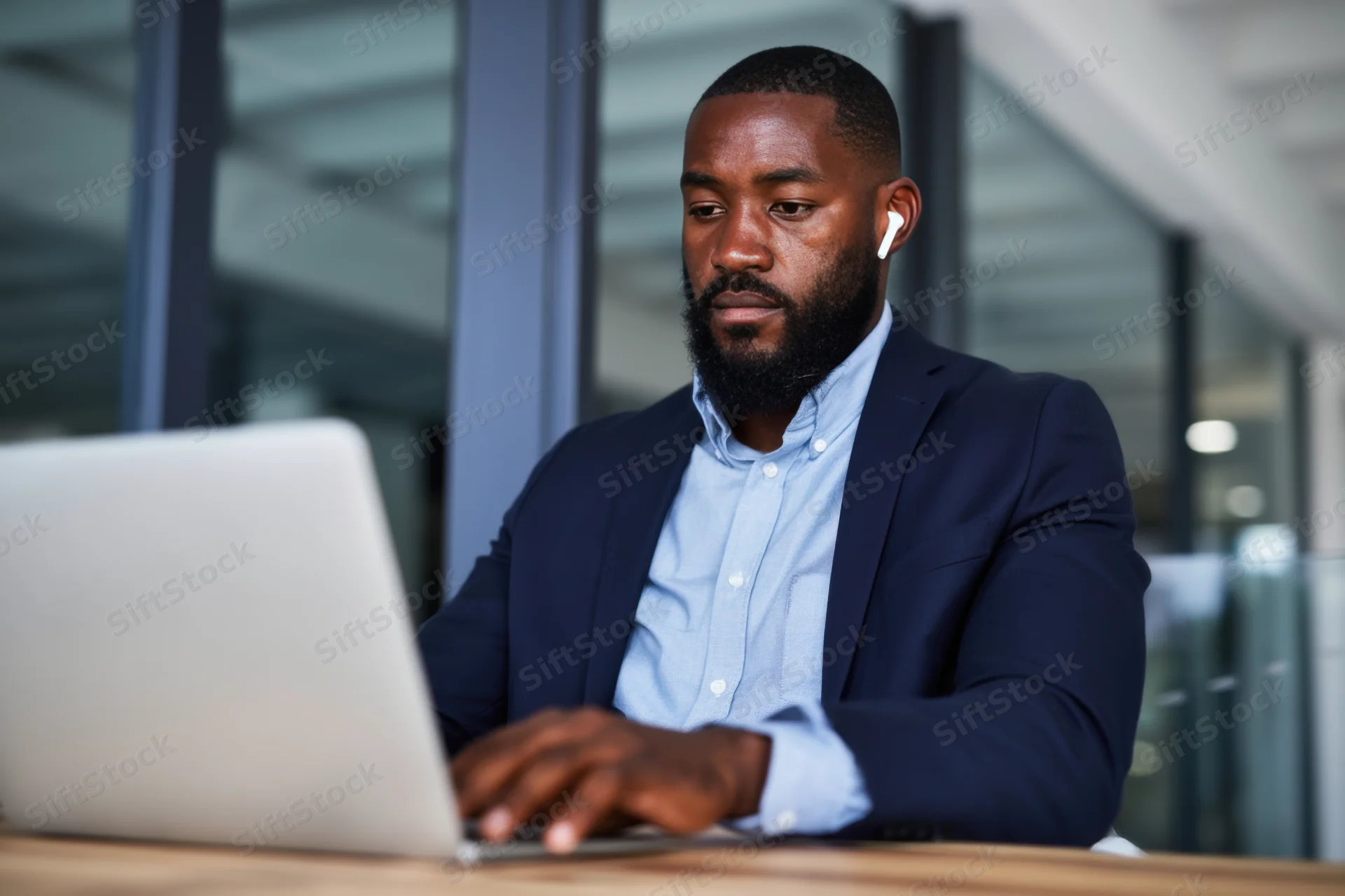 Man in navy blazer and blue shirt typing on laptop at office desk with earbuds in, focused on screen