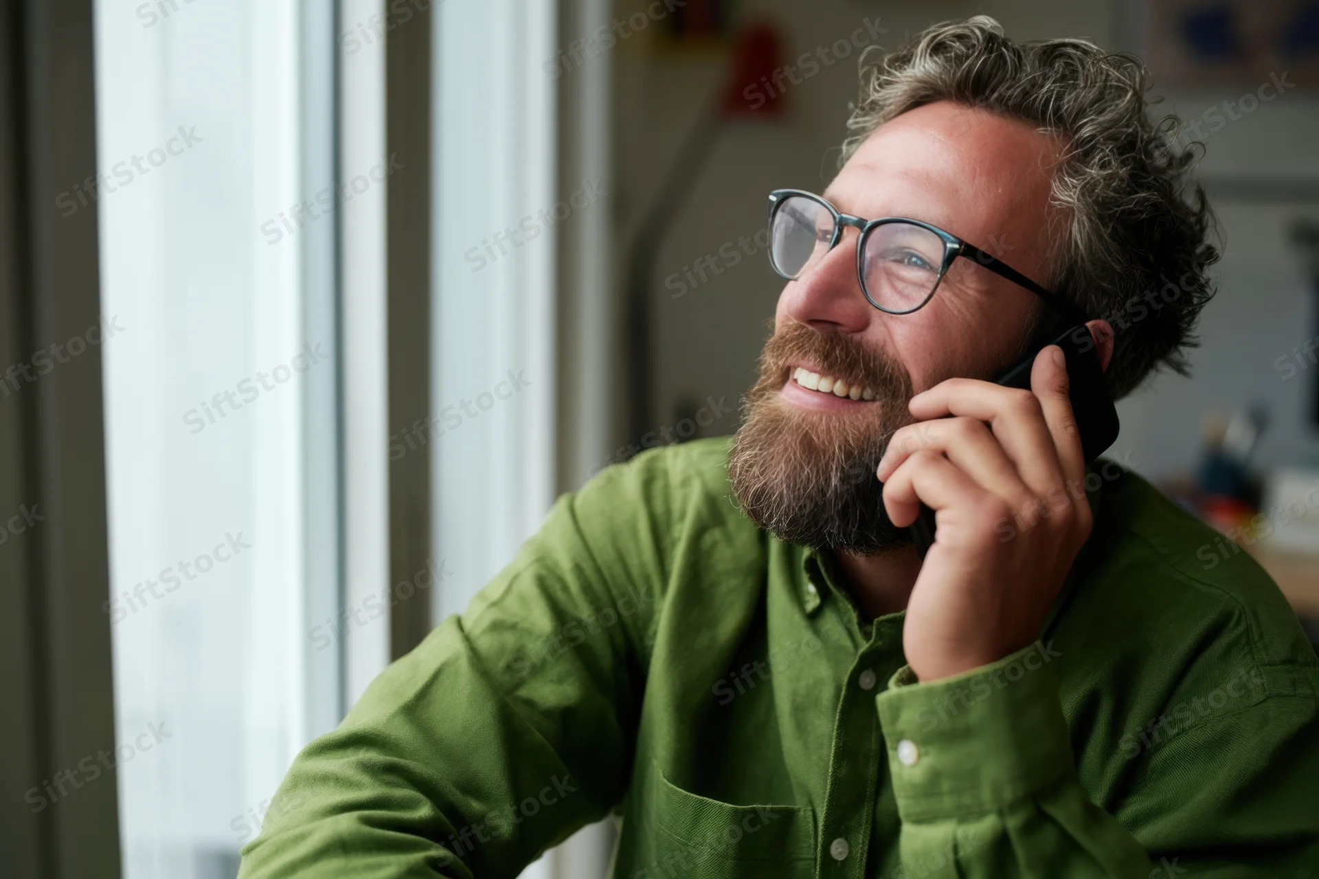 Bearded man in green button-down shirt smiling while talking on phone near window in office