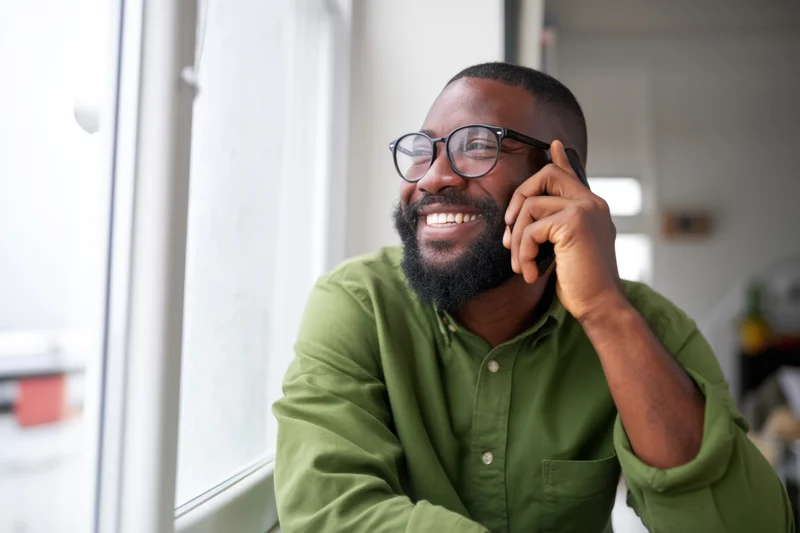 Bearded man with glasses smiling while talking on phone near window indoors