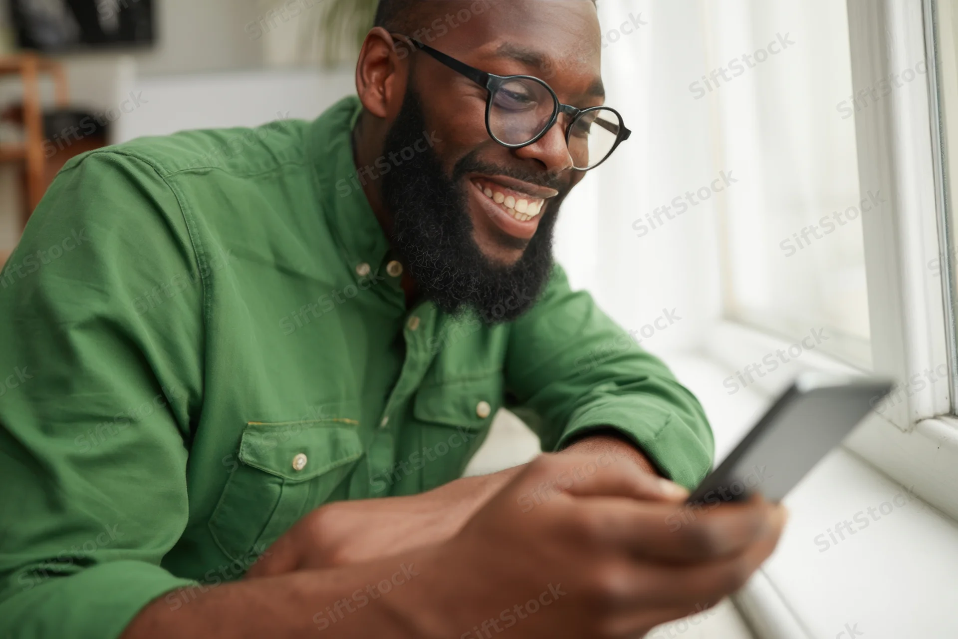 Bearded man in green shirt smiling while using smartphone near window at home