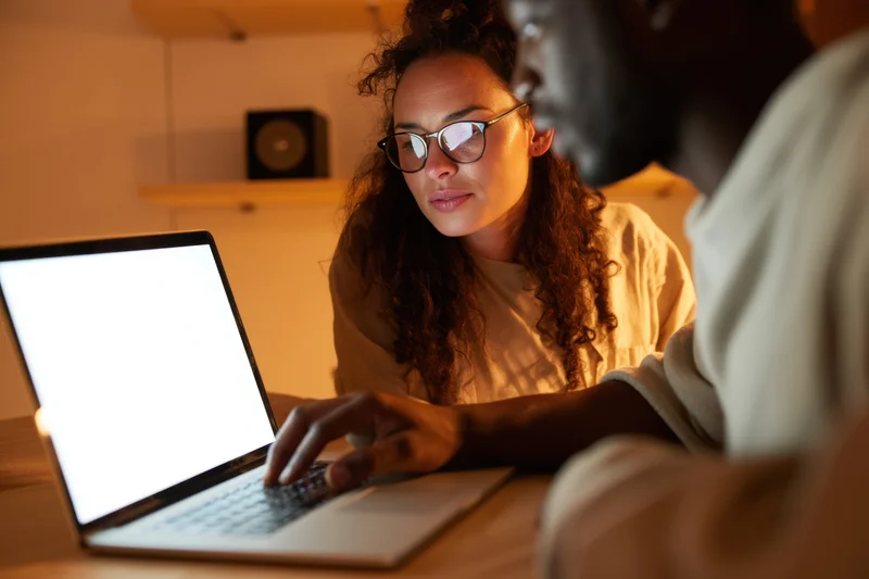 Woman and man collaborating on laptop at wooden table in warm-lit home office