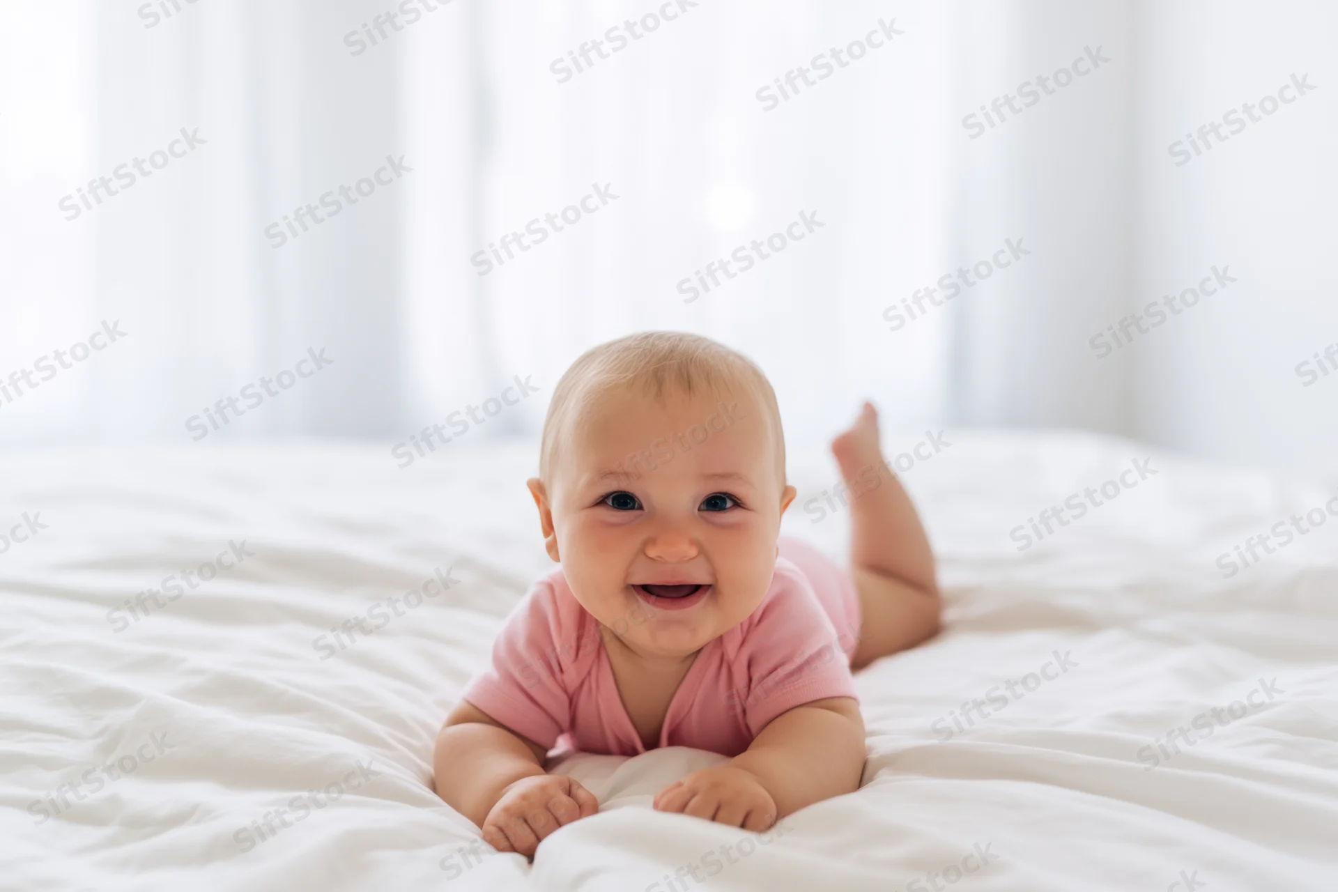 Smiling baby in pink onesie lying on white bed crawling with feet raised