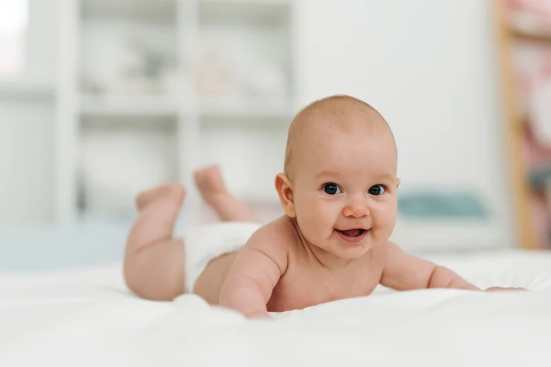 Baby lying on stomach on white surface smiling with diaper on