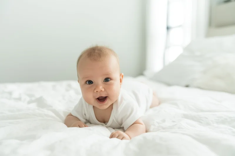Baby lying on stomach on white bed propped on elbows smiling