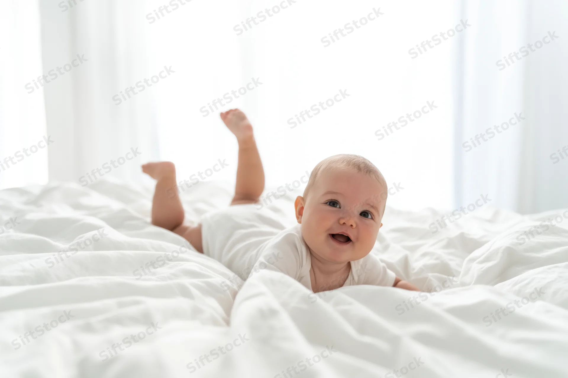 Smiling baby lying on white bed in onesie with legs raised near window