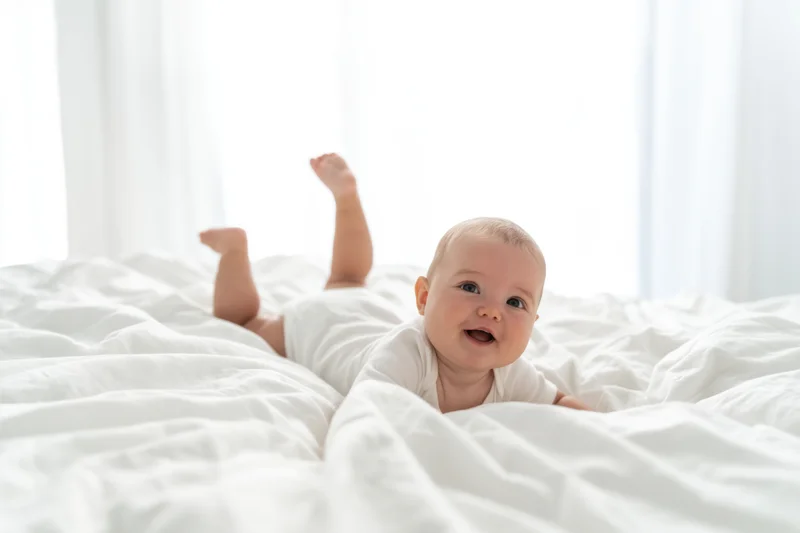 Smiling baby lying on white bed in onesie with legs raised near window