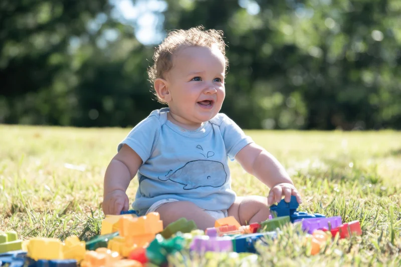 Smiling baby sitting on grassy lawn surrounded by colorful building blocks during daytime playtime