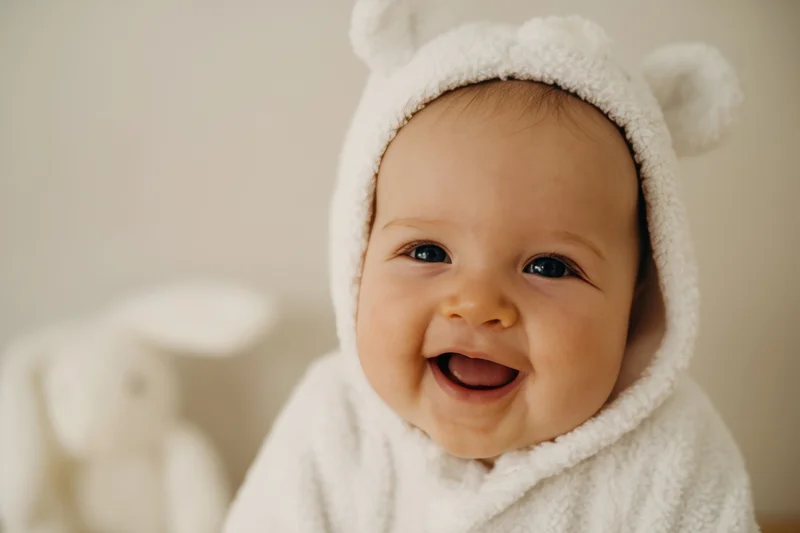 Baby wearing white bear-ear hooded towel smiling with open mouth indoors