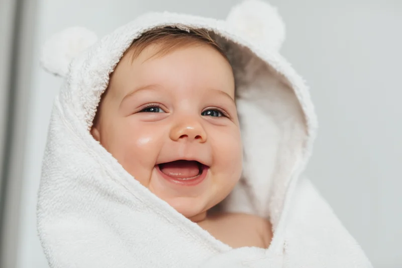 Baby wrapped in a fluffy white hooded towel smiling broadly indoors