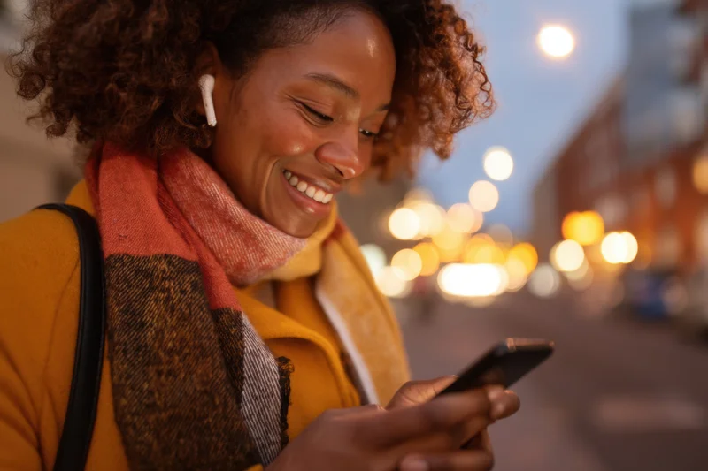 Woman smiling while using smartphone on city street at twilight