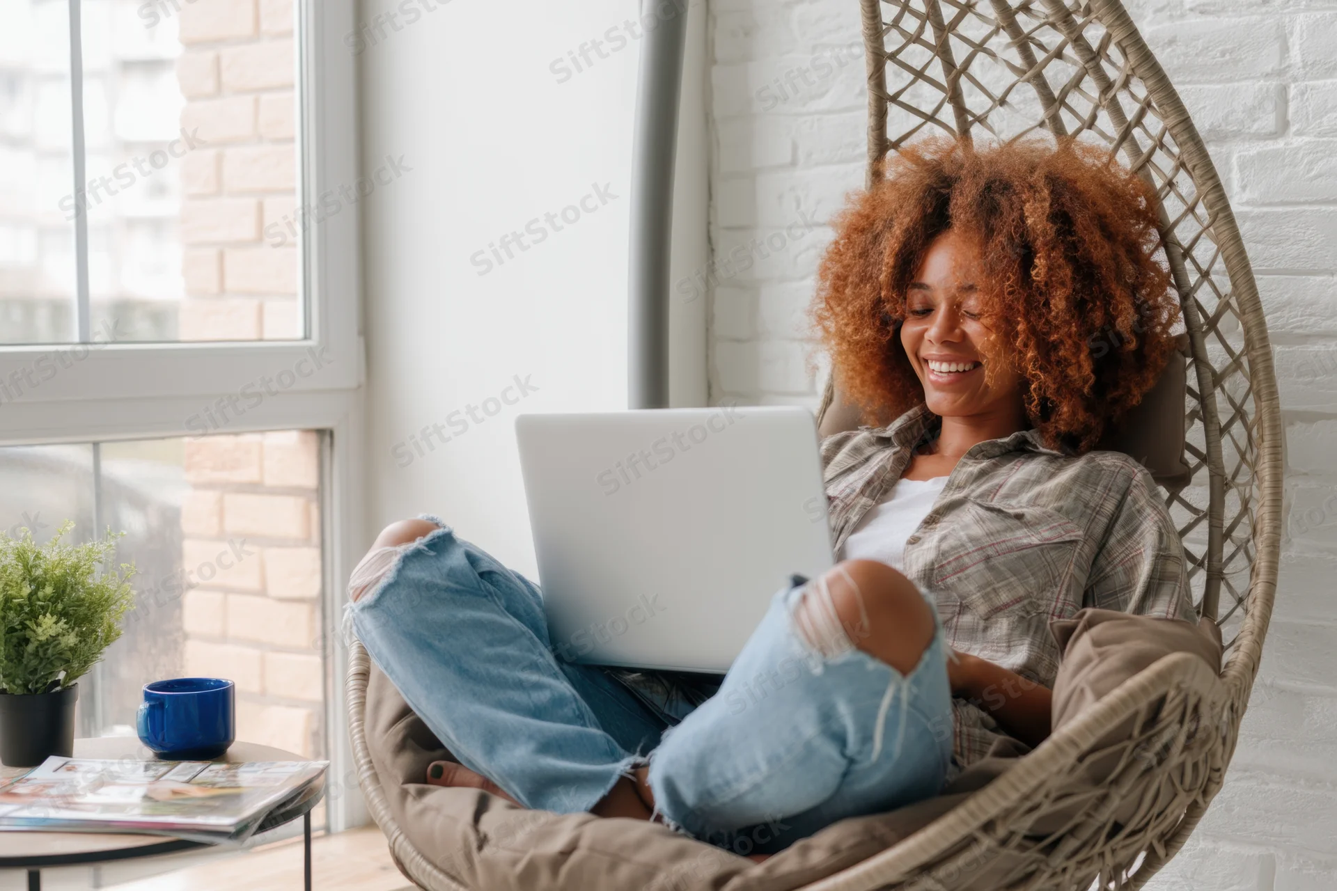 Woman smiling while using laptop cross-legged in hanging wicker chair at home