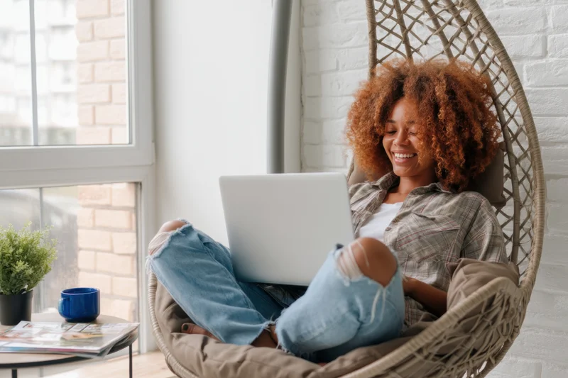 Woman smiling while using laptop cross-legged in hanging wicker chair at home