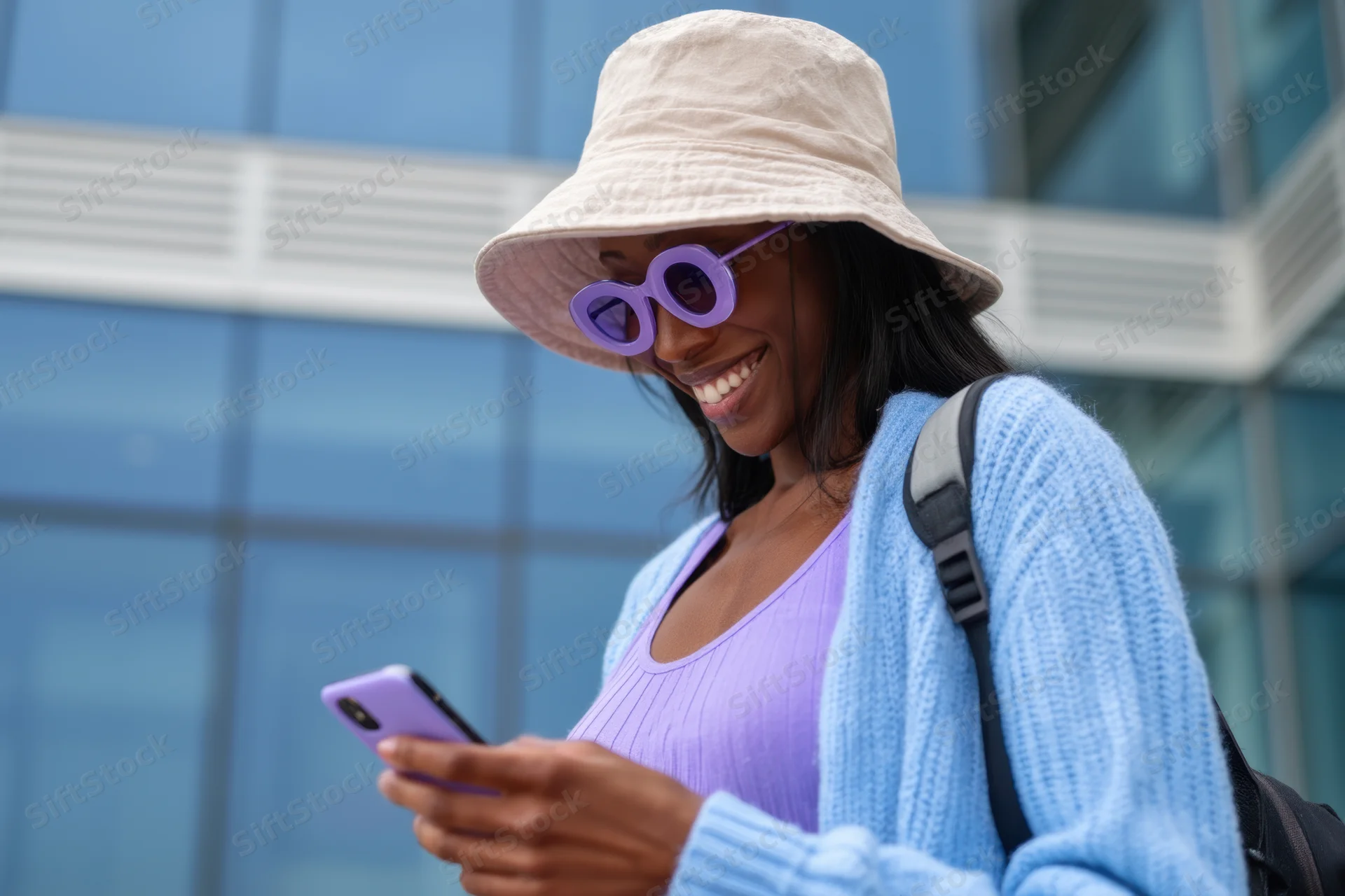 Smiling woman using smartphone outdoors in city wearing bucket hat and sunglasses