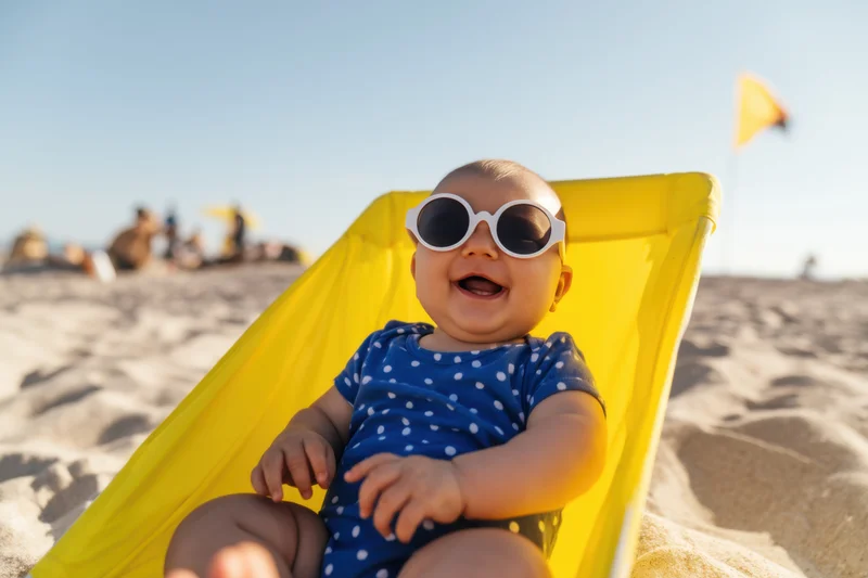 Baby in blue polka-dot onesie wearing white sunglasses laughing in yellow beach chair on sandy shore