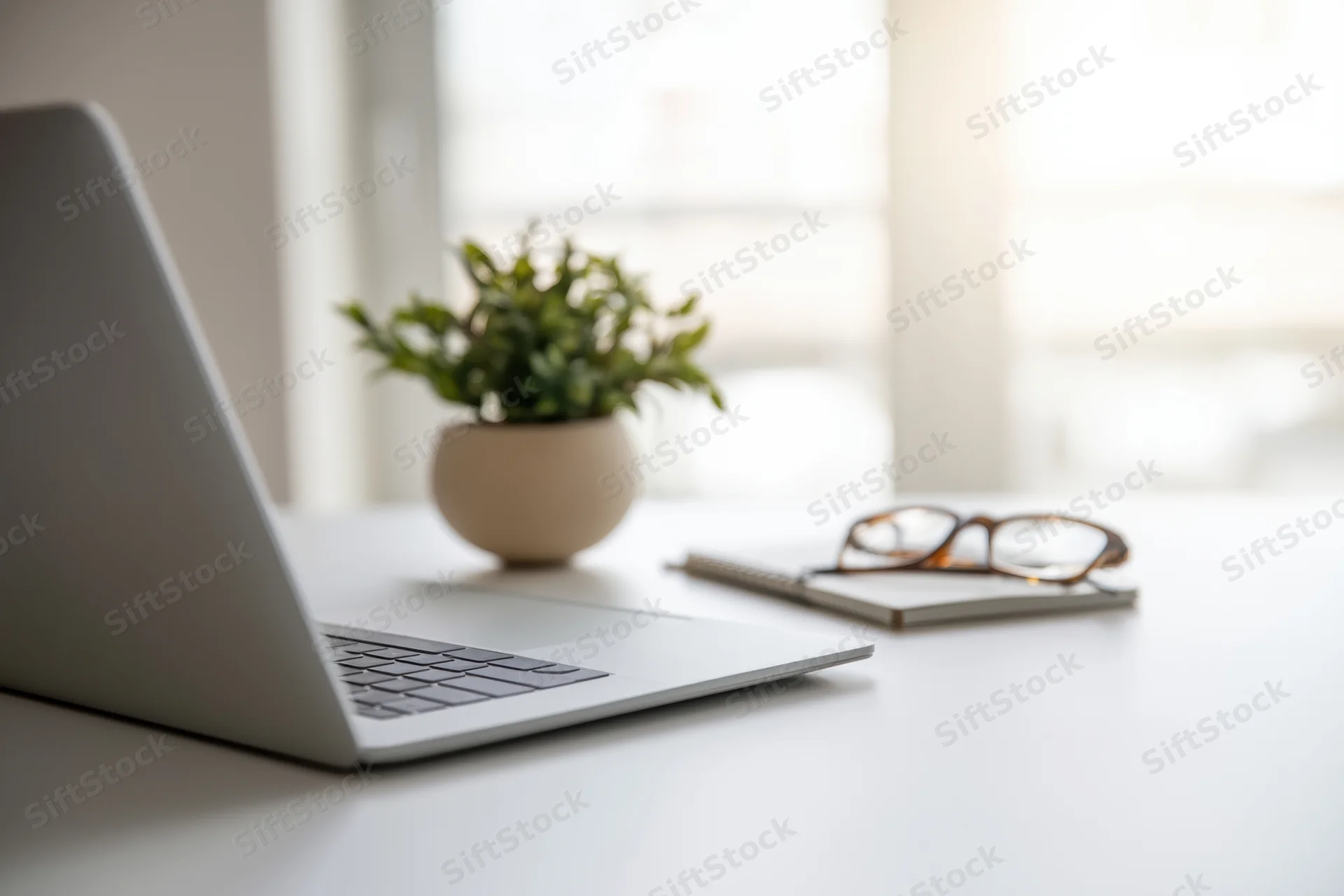 Silver laptop open on white desk beside small potted plant and notebook with glasses, modern office workspace setup