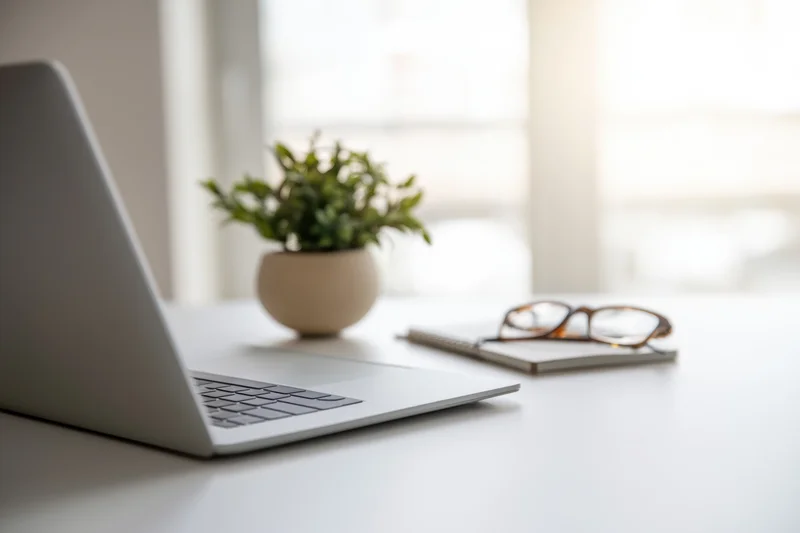 Silver laptop open on white desk beside small potted plant and notebook with glasses, modern office workspace setup