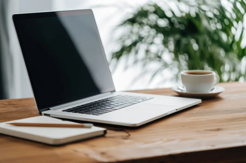 Open silver laptop on wooden desk with coffee cup and notebook beside a green plant