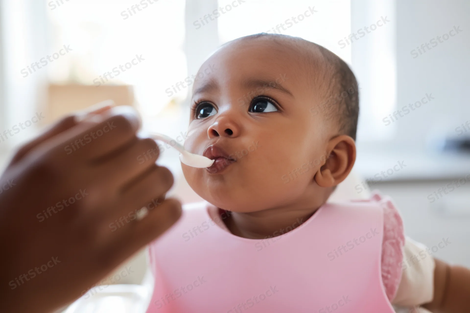 Baby in pink bib being fed with spoon by caregiver indoors