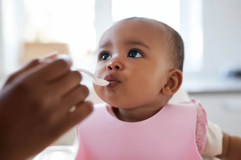 Baby in pink bib being fed with spoon by caregiver indoors