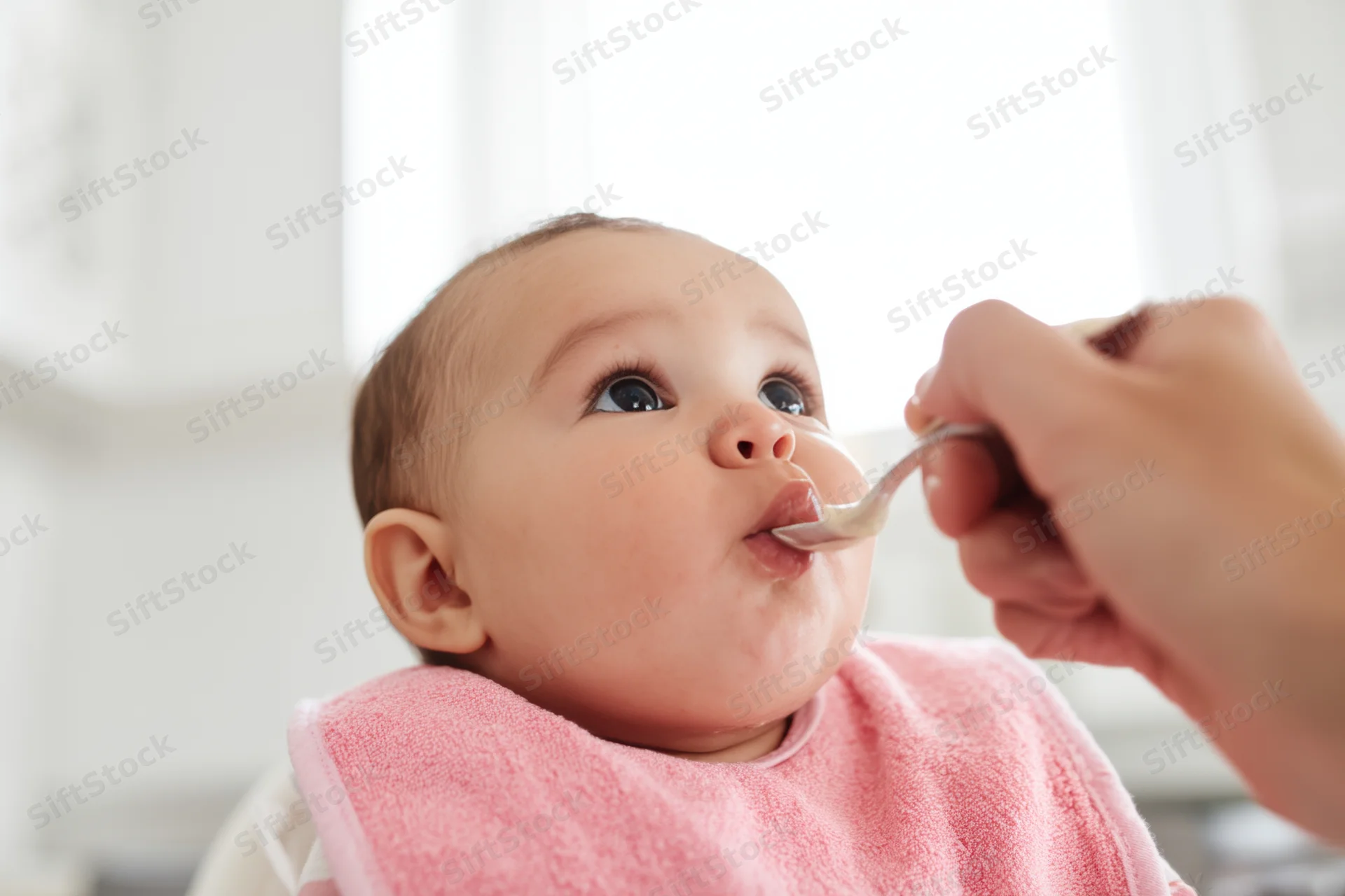 Baby in pink bib being fed with spoon by caregiver in kitchen setting