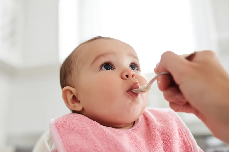 Baby in pink bib being fed with spoon by caregiver in kitchen setting