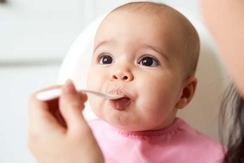 Baby in pink bib eating from spoon held by caregiver in home setting