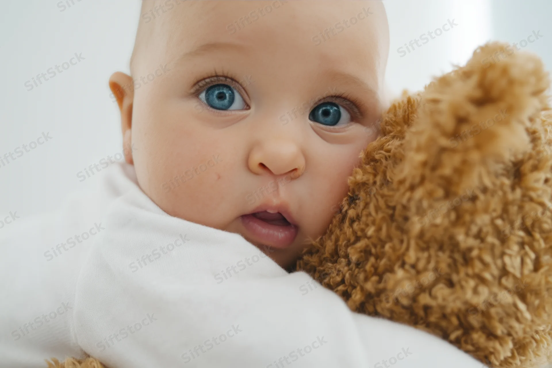 Baby hugging a fluffy brown teddy bear while wrapped in a white blanket