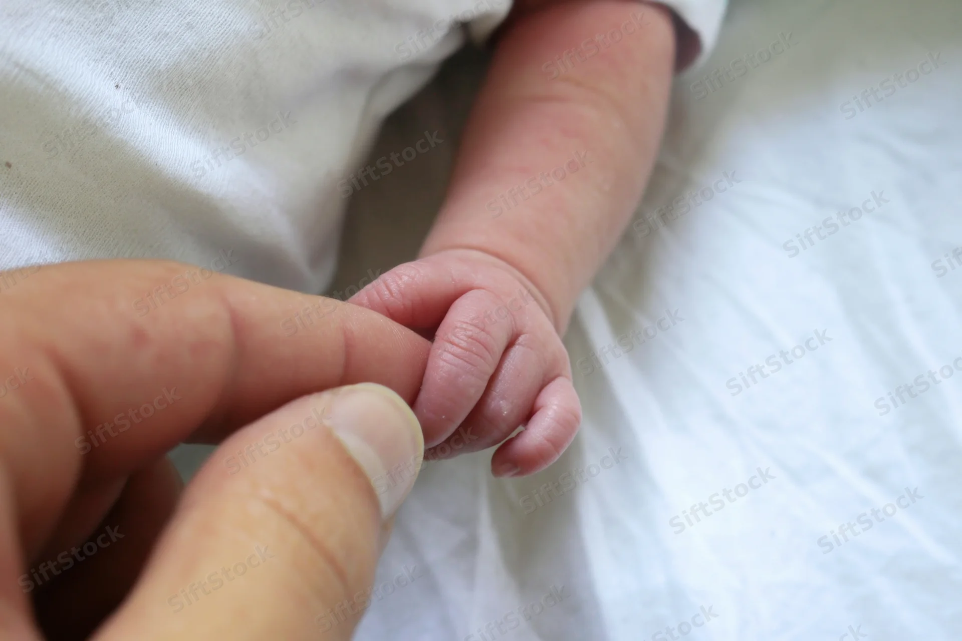 Newborn baby's tiny hand holding an adult finger on a white surface