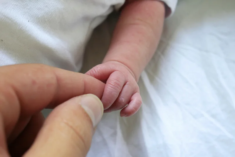 Newborn baby's tiny hand holding an adult finger on a white surface