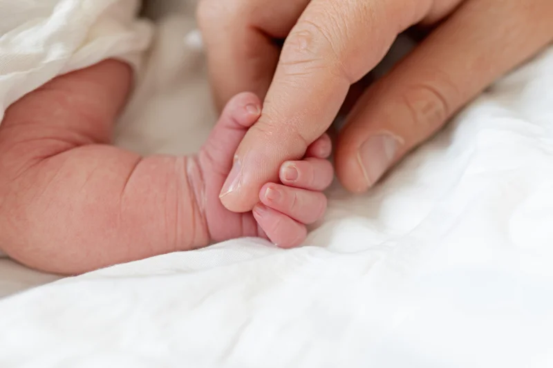 Newborn’s tiny hand grasping an adult finger on a white blanket