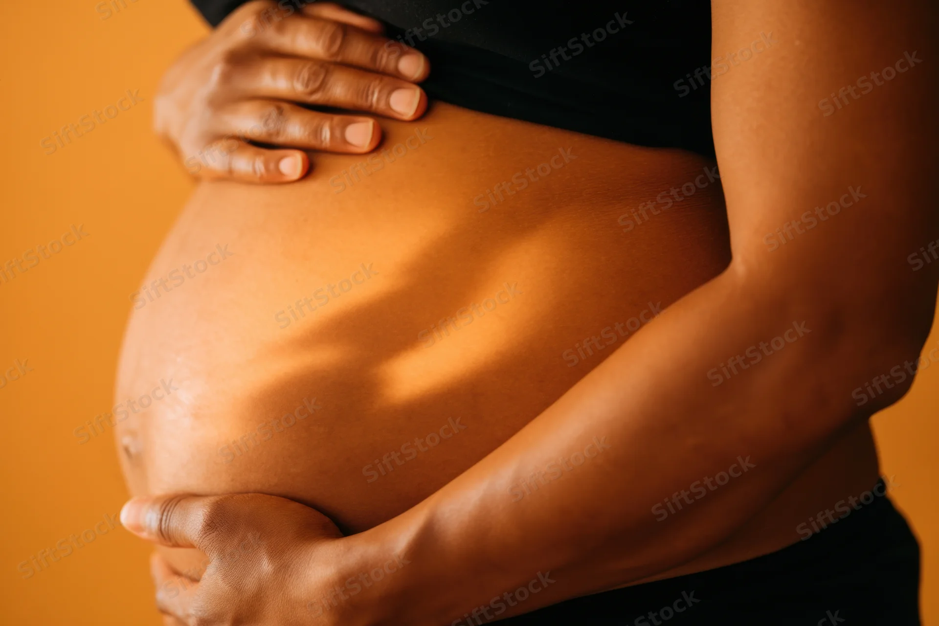 Pregnant woman cradling her belly with hands in warm light against orange background