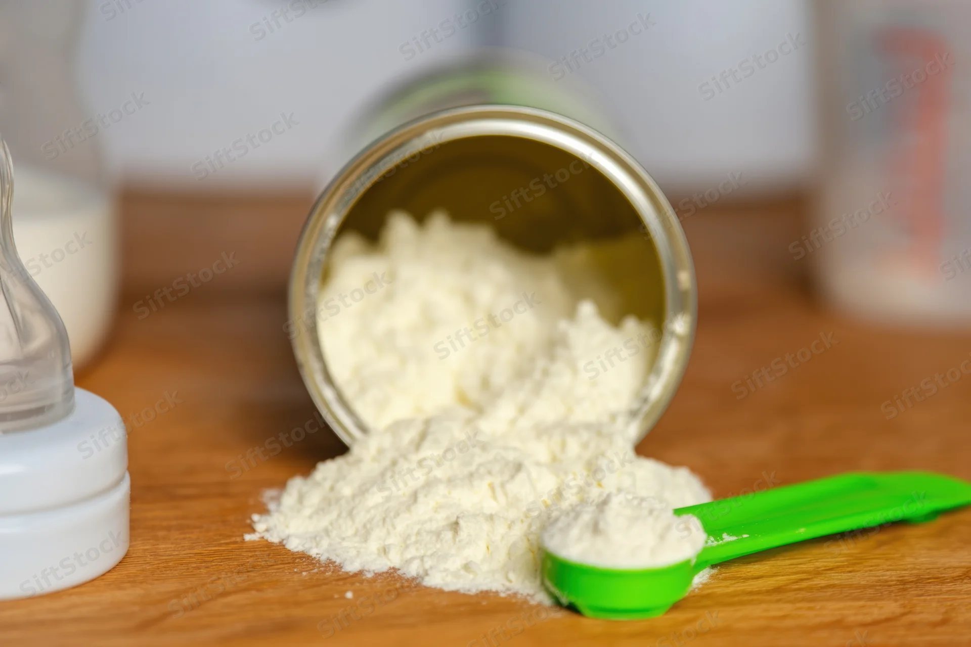 Spilled baby formula powder on wooden table with green scoop and bottle nearby