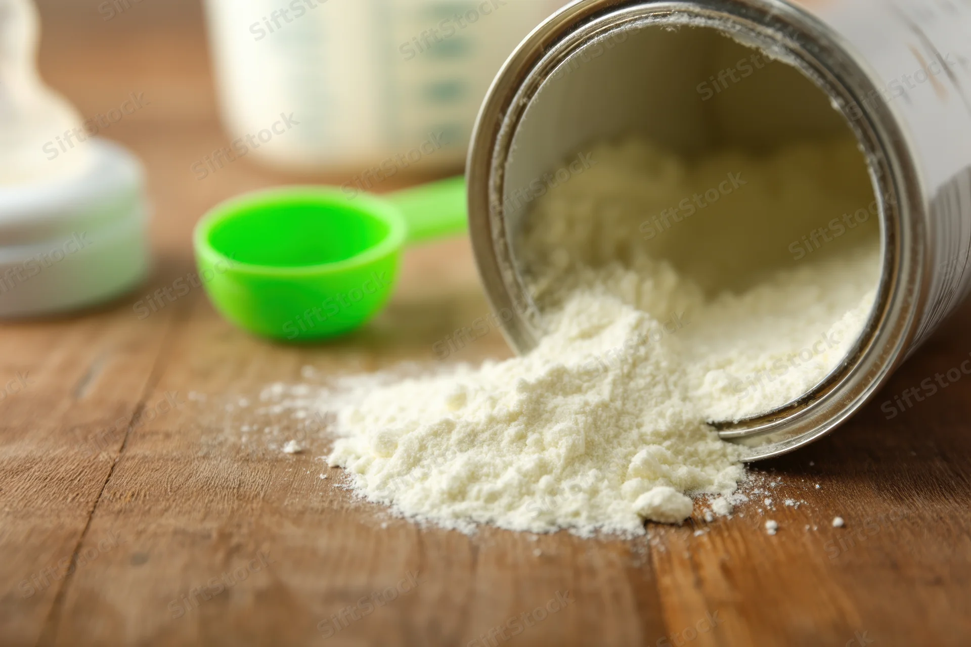 Spilled baby formula powder from a container on a wooden table with a green scoop nearby