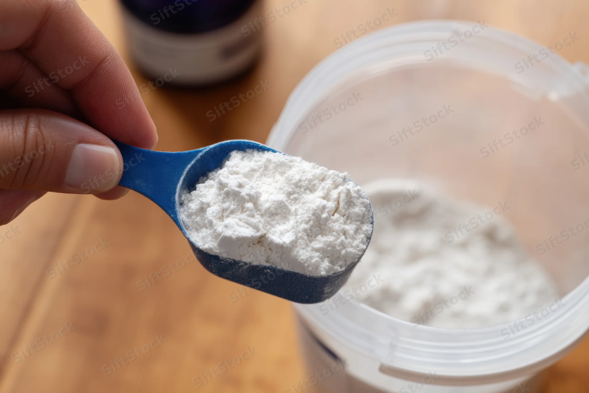 Hand holding blue scoop filled with white protein powder over plastic container on wooden table