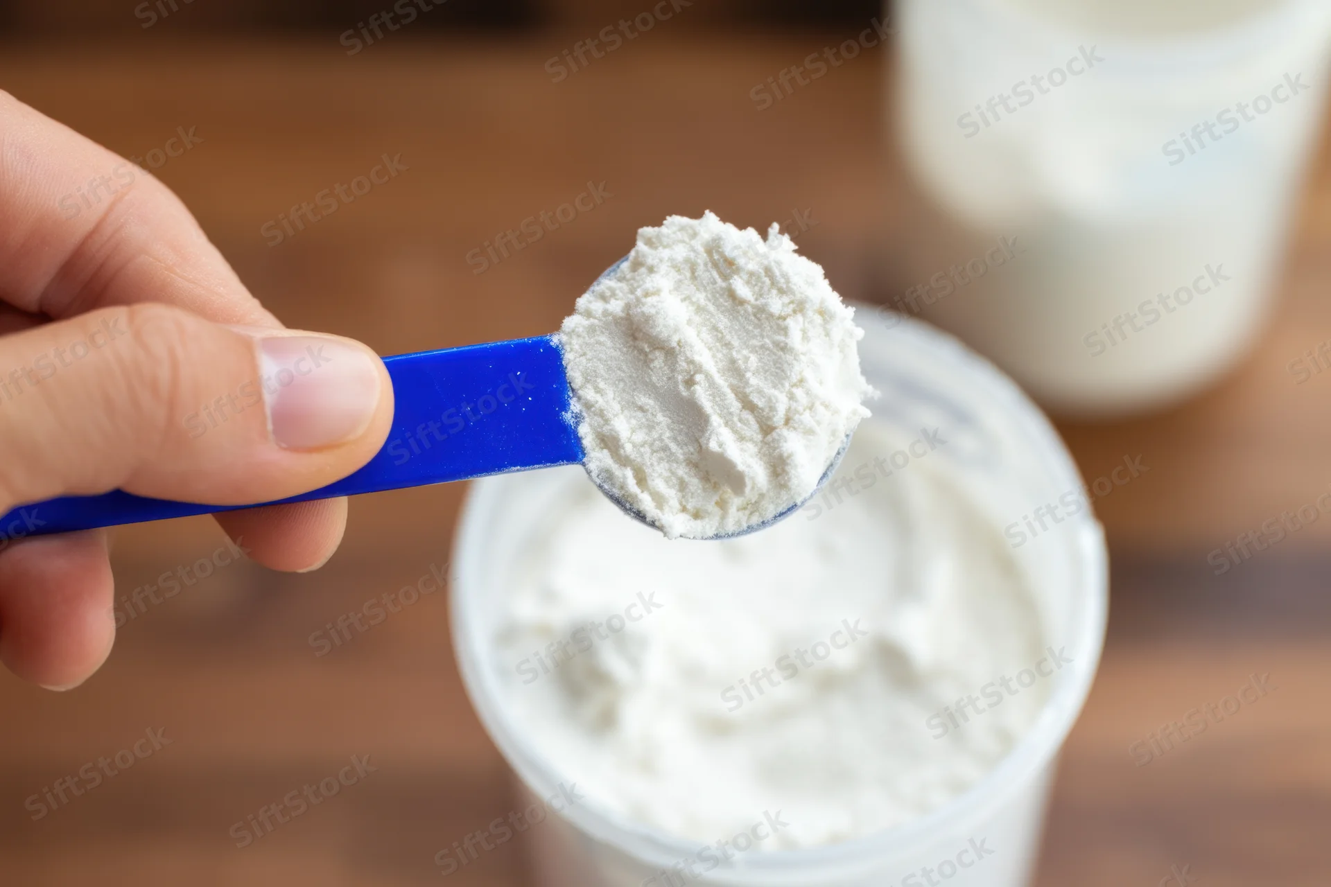 Hand holding blue scoop filled with white protein powder over container on wooden surface
