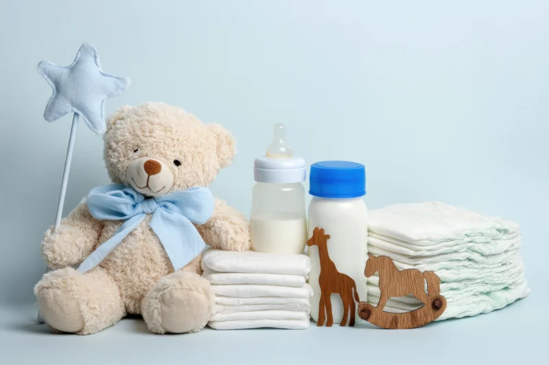 Plush teddy bear with blue bow and star wand beside baby bottles, diapers, and wooden toys on light blue background