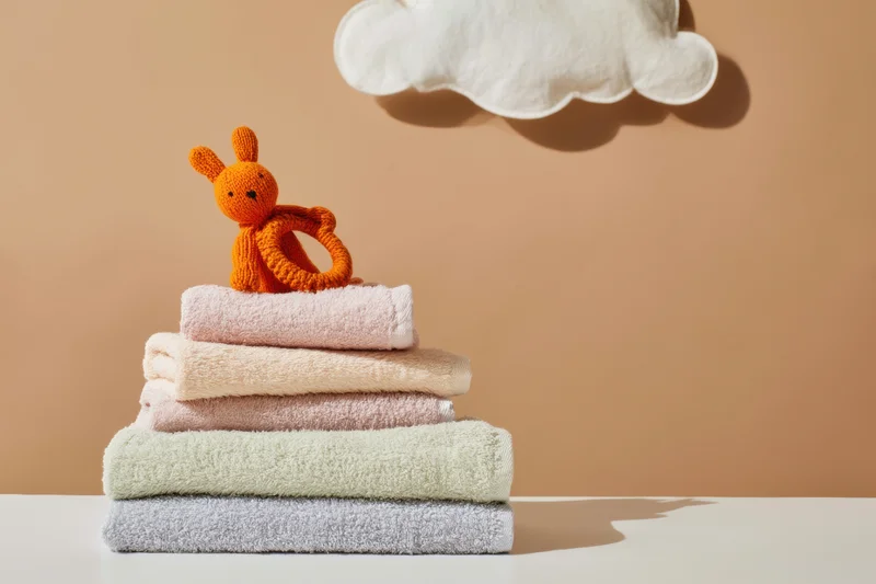 Stack of pastel folded towels with knitted orange bunny teether on top near cloud decor on neutral background