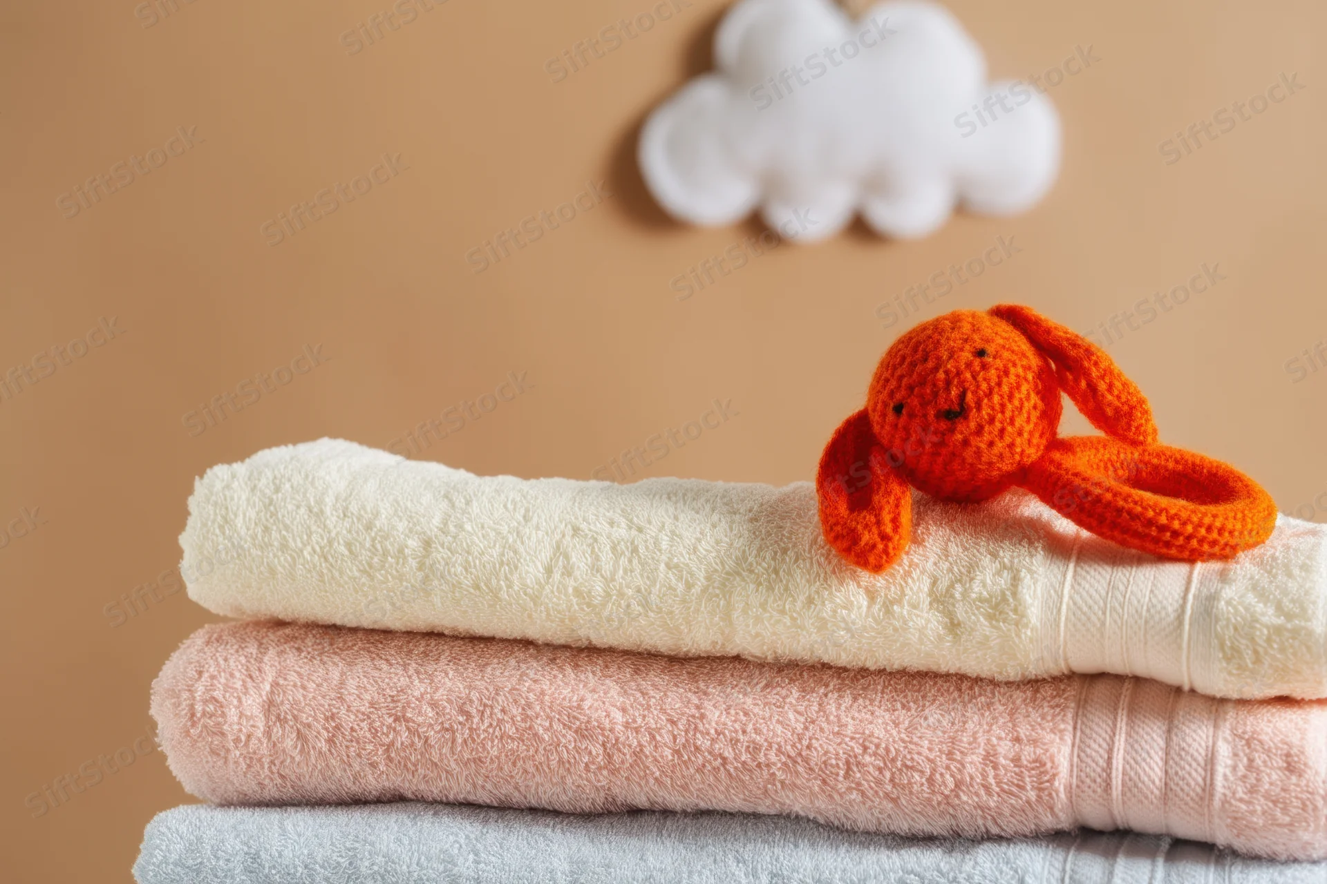 Stack of pastel folded towels with crocheted orange rabbit toy on top near cloud decor on beige wall