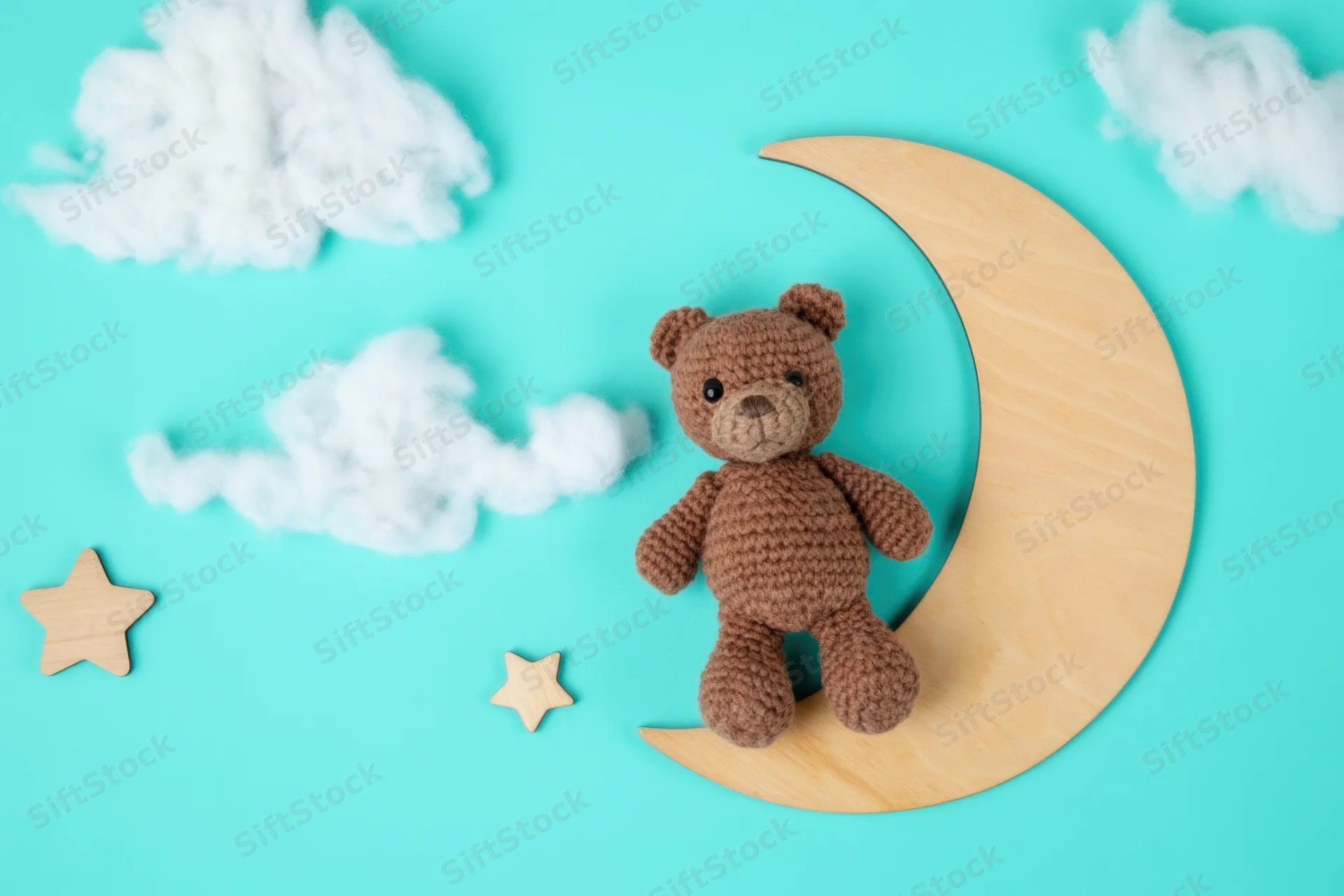 Crocheted brown teddy bear sitting on wooden crescent moon against turquoise background with clouds and stars