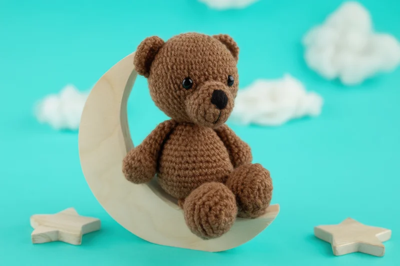 Crocheted brown teddy bear sitting on a wooden crescent moon with stars and clouds on turquoise background