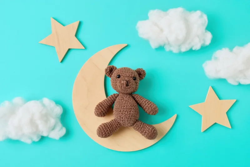 Crocheted brown teddy bear sitting on wooden crescent moon surrounded by stars and clouds on turquoise background