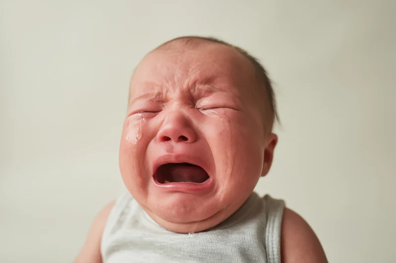 Crying baby with eyes squeezed shut and mouth open in a wail, wearing a light gray sleeveless top against a plain background